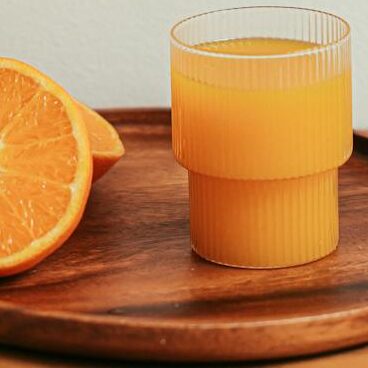 A glass of fresh orange juice with oranges on a wooden tray, showcasing a refreshing citrus beverage.